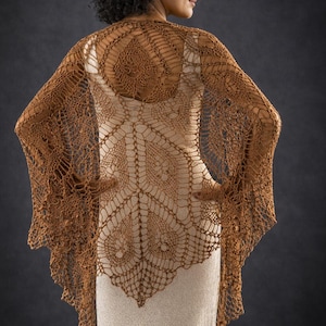 Puede incluir: Un chal de ganchillo marrón con un patrón calado y detallado. El chal está sobre un vestido color crema. El intrincado diseño presenta formas circulares y geométricas, creando un aspecto delicado y elegante. La textura del chal es visible.