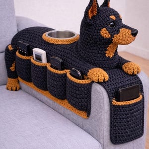 Pode incluir: Organizador de apoio de braço de sofá em forma de cão de crochê em preto e castanho. Possui um porta-copos, vários bolsos para controles remotos e telefones e um design decorativo. Um item de decoração para casa único e funcional.