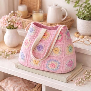 May include: A pink crocheted tote bag with a floral pattern. The bag features a light pink body with colorful floral squares and cream-colored straps. The bag is sitting on a stack of books.