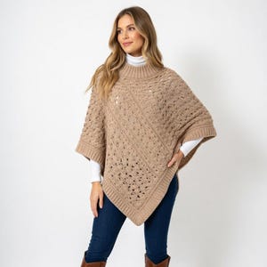 Könnte beinhalten: Ein beigefarbener Strickponcho mit hohem Rippkragen und kurzen Ärmeln. Der Poncho hat ein strukturiertes, offen gewebtes Design und einen quadratischen Saum. Das Model trägt einen weißen Rollkragenpullover darunter und blaue Jeans.
