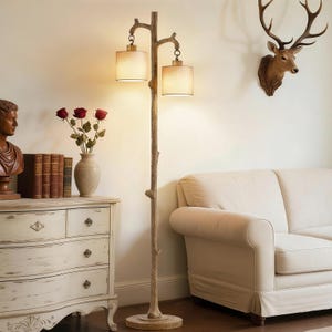 May include: A rustic floor lamp with a tree branch design. The lamp has two beige fabric shades and a textured, brown base. The lamp is next to a white sofa and a chest of drawers. A deer head decorates the wall.