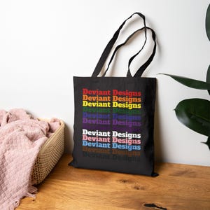 Może przedstawiać: Czarna torba typu tote z dwoma paskami na ramię, na której nadrukowano tekst "Deviant Designs" w kilku rzędach, każdy w innym kolorze, w tym czerwonym, pomarańczowym, żółtym, zielonym, niebieskim, fioletowym, białym, różowym, jasnoniebieskim, brązowym i szarym.