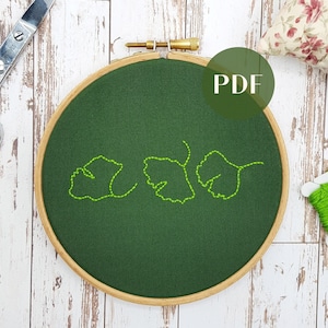 May include: A green embroidery hoop with a design of three green leaves stitched in a line. The hoop is made of wood and has a natural finish. The background is a white wooden surface. The text "PDF" is visible in a green circle.