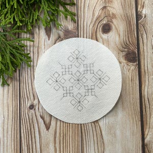 May include: A circular, white coaster featuring a grey geometric snowflake pattern. The coaster is made of a textured material and rests on a wooden surface, with green foliage in the background.