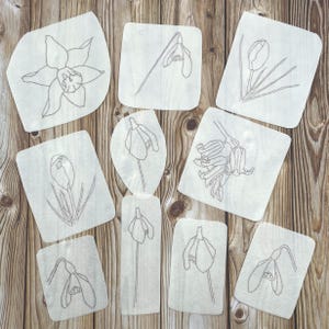 May include: A selection of white fabric patches featuring embroidered line drawings of spring flowers, such as daffodils, snowdrops, and tulips. The patches are displayed on a wooden surface, highlighting the intricate floral designs.