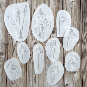 May include: A selection of white fabric patches featuring embroidered snowdrop flower designs. The patches are in various sizes and shapes, with grey stitching detailing the flowers and stems. The background is a wooden surface.