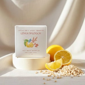 Op de afbeelding: Een doosje "Lemon Splendor" ambachtelijke thee met natuurlijke ingrediënten, met een citroenillustratie. De afbeelding bevat verse citroenen, een citroenpartje en een hoop gerst. De doos staat op een wit voetstuk.