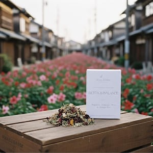 Puede incluir: Una caja de madera exhibe una caja blanca y un montón de hojas de té sueltas. La caja dice "Detox & Balance" con texto adicional. Flores rojas y rosas bordean una calle con edificios tradicionales al fondo.