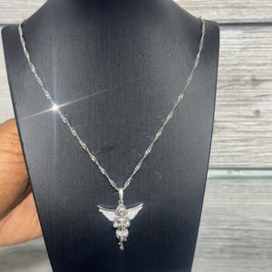 K&ouml;nnte beinhalten: Eine silberfarbene Halskette mit einem Anh&auml;nger in Form eines Engels. Der Engel hat ausgebreitete Fl&uuml;gel und ist mit kleinen, funkelnden Steinen besetzt. Die Halskette wird auf einem schwarzen Schmuckst&auml;nder vor hellem Holzdekor pr&auml;sentiert.