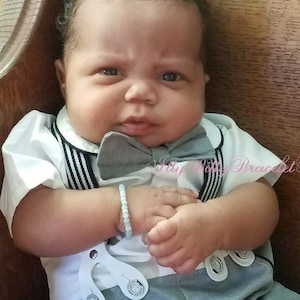 May include: A baby boy wearing a gray and white outfit with a bow tie. He is sitting on a wooden bench. The text "My Baby Bracelet Boutique.com" is visible in the background.