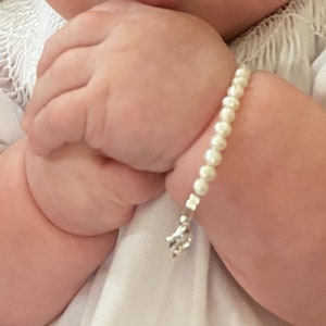 Puede incluir: Una delicada pulsera de perlas con un dije plateado adorna la mu&ntilde;eca de un beb&eacute;. La pulsera est&aacute; hecha de peque&ntilde;as perlas blancas redondas. El dije es un peque&ntilde;o dise&ntilde;o plateado.
