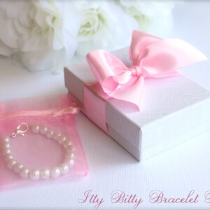 Puede incluir: Un brazalete de perlas rosas en una bolsa de organza rosa con un cierre plateado. El brazalete est&aacute; empaquetado en una caja de regalo blanca con un lazo de sat&eacute;n rosa.