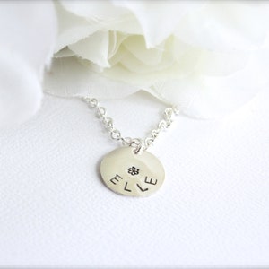 May include: A silver necklace with a round pendant that says "ELLE" with a small flower design in the center.