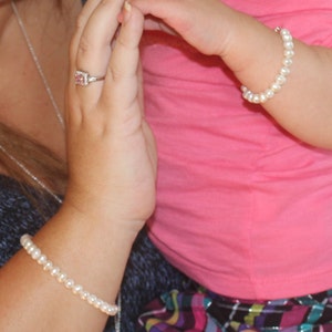 May include: Two people wearing pearl bracelets. The bracelet on the adult's wrist is a simple strand of pearls, while the bracelet on the child's wrist is a single strand of pearls with a small, pink stone accent.