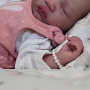 Puede incluir: Un beb&eacute; dormido con un conjunto rosa con lunares blancos y un body blanco. La mu&ntilde;eca del beb&eacute; tiene una pulsera de perlas. El beb&eacute; est&aacute; acostado sobre una manta suave estampada.