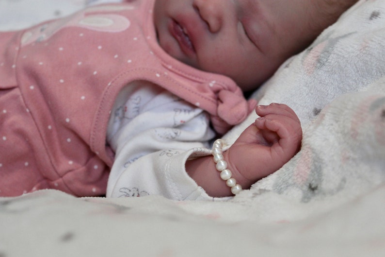 Puede incluir: Un beb&eacute; dormido con un conjunto rosa con lunares blancos y un body blanco. La mu&ntilde;eca del beb&eacute; tiene una pulsera de perlas. El beb&eacute; est&aacute; acostado sobre una manta suave estampada.