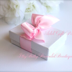 Puede incluir: Una peque&ntilde;a caja de regalo blanca atada con una cinta y un lazo de sat&eacute;n rosa claro. La caja tiene el texto "Add Gift Packaging" y "Itty Billy Bracelet Boutique.com" en escritura rosa.