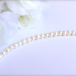 May include: A delicate pearl necklace with small, round, off-white pearls. The necklace is strung together with a silver clasp. White flowers are in the background, creating a soft, elegant aesthetic. The pearls are approximately 0.2 inches in diameter.