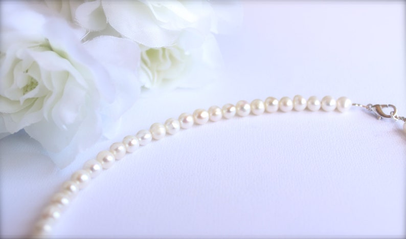 May include: A delicate pearl necklace with small, round, off-white pearls. The necklace is strung together with a silver clasp. White flowers are in the background, creating a soft, elegant aesthetic. The pearls are approximately 0.2 inches in diameter.