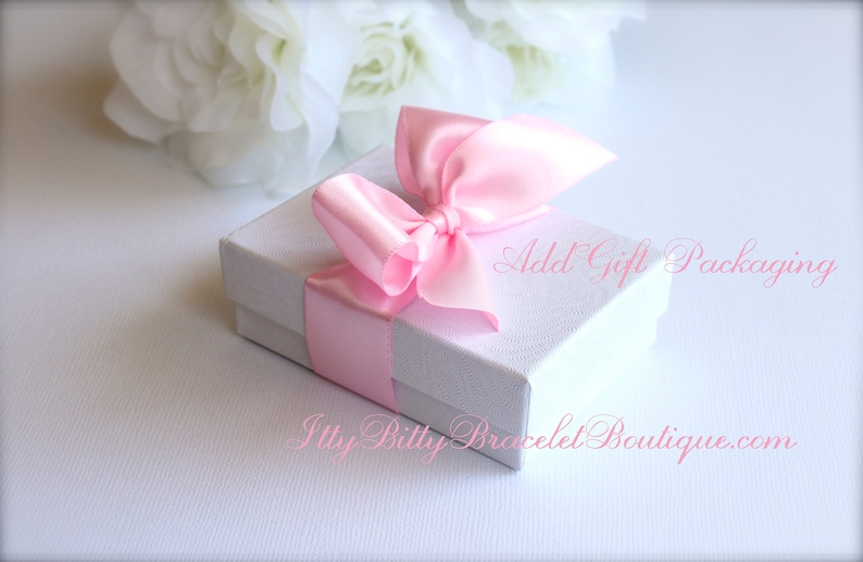 May include: A small, white gift box with a pink satin ribbon and bow. The box is tied with a pink ribbon and bow. The text "Add Gift Packaging" is written on the box. White flowers are in the background.