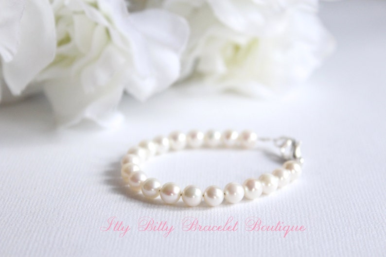 Puede incluir: Un delicado brazalete con una hilera de perlas redondas de color blanco cremoso. El brazalete se asegura con un cierre plateado. El texto "Itty Billy Bracelet Boutique" est&aacute; en rosa.