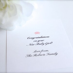 Puede incluir: Tarjeta blanca con el texto &laquo;&iexcl;Felicidades por tu nueva ni&ntilde;a!&raquo; en una fuente elegante, coronada por una corona rosa. Se ve una flor blanca.