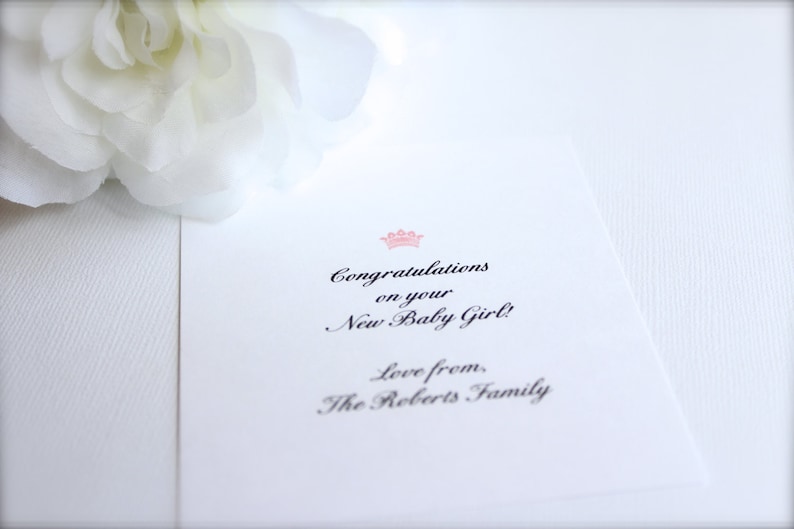 Puede incluir: Tarjeta blanca con el texto &laquo;&iexcl;Felicidades por tu nueva ni&ntilde;a!&raquo; en una fuente elegante, coronada por una corona rosa. Se ve una flor blanca.