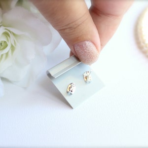 May include: A pair of small, silver-colored stud earrings on a light blue display card. The earrings have a rounded, organic shape. The background includes white flowers and a string of pearls.