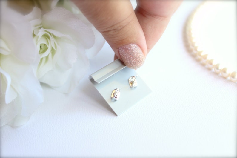 May include: A pair of small, silver-colored stud earrings on a light blue display card. The earrings have a rounded, organic shape. The background includes white flowers and a string of pearls.