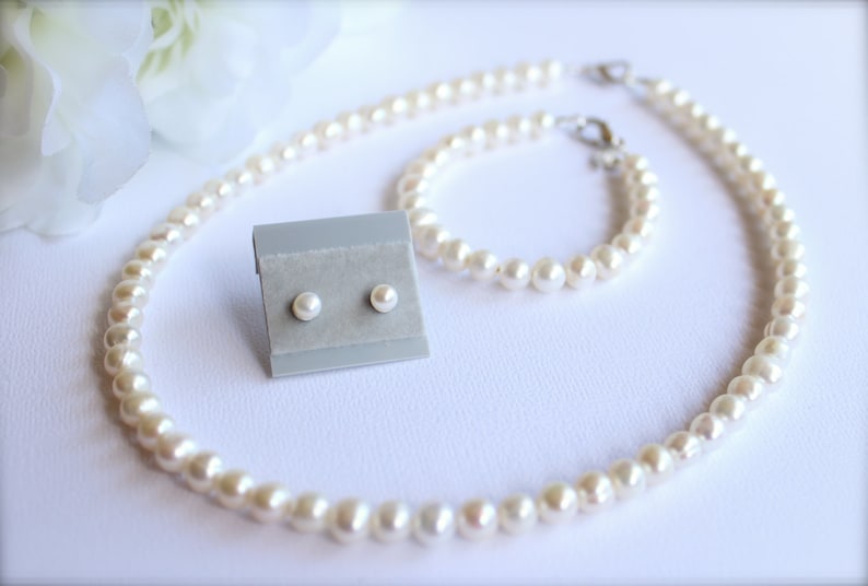May include: A pearl jewelry set including a necklace, bracelet, and stud earrings. The necklace and bracelet are strung with small, round pearls. The earrings are displayed on a gray card. The set is on a white surface.