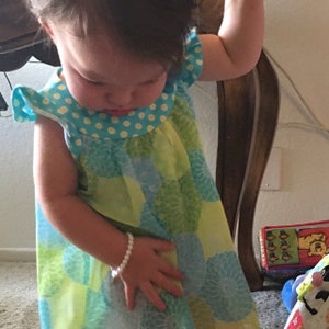 Puede incluir: Una ni&ntilde;a peque&ntilde;a con un vestido sin mangas con un estampado floral azul y verde. El vestido tiene un cuello azul con lunares y una pulsera de perlas blancas en la mu&ntilde;eca.