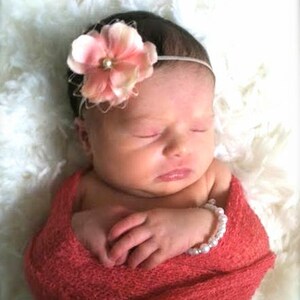 Puede incluir: Un beb&eacute; durmiendo con una diadema de flores rosas y un brazalete de perlas blancas envuelto en una manta de color coral.