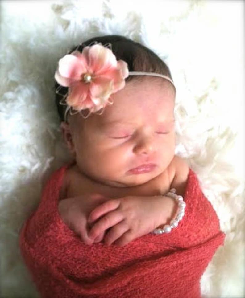 Puede incluir: Un beb&eacute; durmiendo con una diadema de flores rosas y un brazalete de perlas blancas envuelto en una manta de color coral.