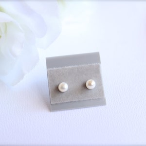 May include: A pair of small, round, pearl earrings displayed on a gray velvet card. The earrings are a creamy white color and are set against a soft, blurred white background with floral accents.