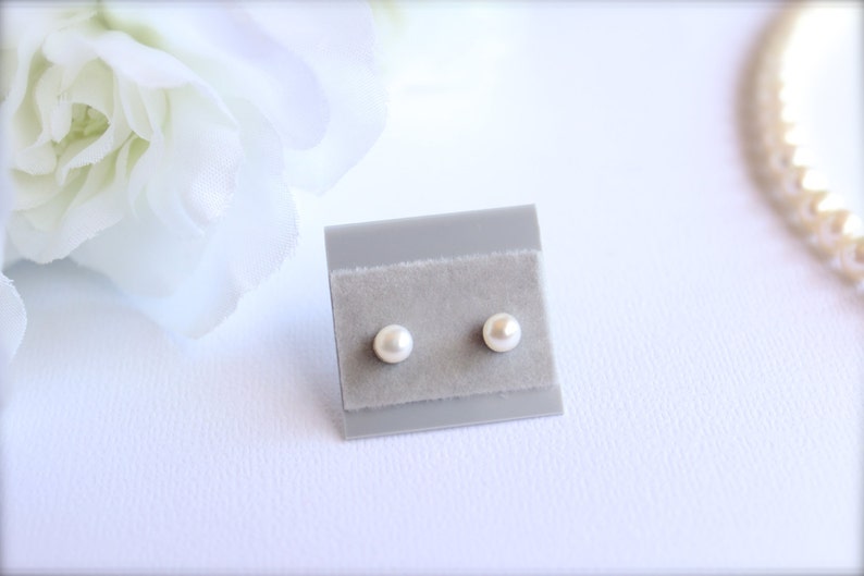 May include: A pair of small, round, pearl earrings displayed on a gray velvet card. The earrings are a creamy white color and are set against a soft, blurred white background with floral accents.