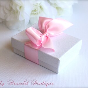 May include: A white gift box with a pink satin ribbon tied in a bow. The box is sitting on a white surface.