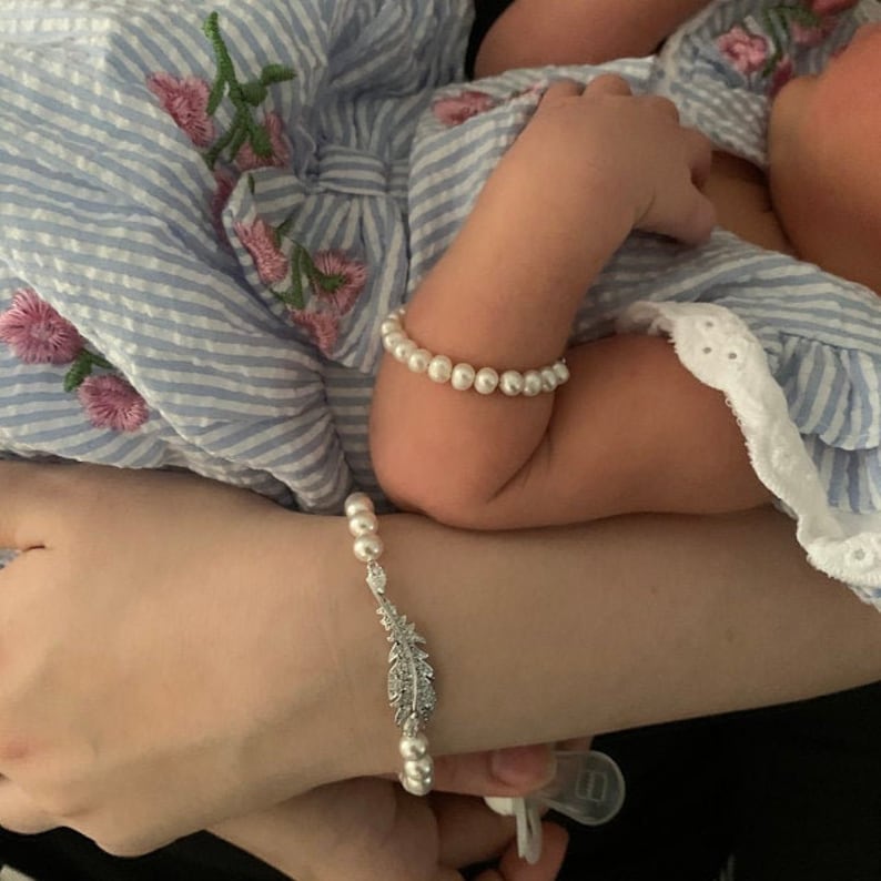 Puede incluir: Un brazo de beb&eacute; adornado con una delicada pulsera de perlas. Una segunda pulsera de perlas rodea la mu&ntilde;eca. El brazo de una madre muestra una pulsera de perlas con un dije de pluma decorativa. El beb&eacute; lleva un vestido a rayas azul y blanco con bordado floral.