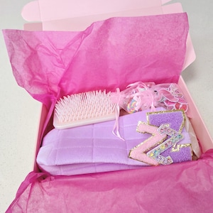 Peut inclure: Une boîte cadeau rose ouverte, remplie de papier de soie rose, d'une brosse violet clair, d'un masque pour les yeux lavande et d'une lettre décorative "Z".