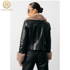 May include: Black leather jacket with a tan fur collar and cuffs. The jacket has a classic design with a tailored fit. The model is wearing sunglasses and black pants. The logo "ELITE LEATHER" is in the upper left corner.
