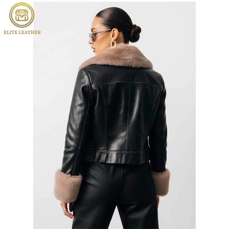 May include: Black leather jacket with a tan fur collar and cuffs. The jacket has a classic design with a tailored fit. The model is wearing sunglasses and black pants. The logo "ELITE LEATHER" is in the upper left corner.