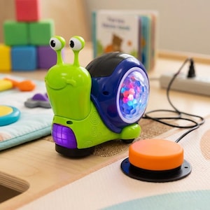 May include: A green and blue toy snail with a light-up shell. The snail has a purple base and an orange button connected by a black wire. The toy is designed for children and sits on a wooden surface.