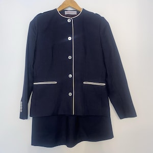 Può includere: Un tailleur gonna blu navy con finiture bianche. La giacca ha un colletto tondo, bottoni bianchi e una gonna abbinata. L'abito è appeso a una gruccia di legno su sfondo bianco. L'abito è uno stile classico.
