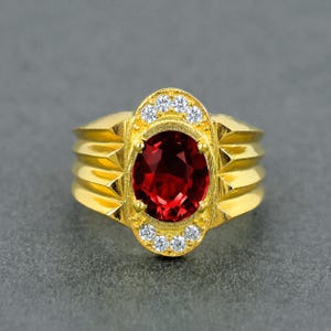 Può includere: Un anello d'oro con una grande pietra preziosa rosso scuro a taglio ovale. L'anello ha un'incastonatura decorativa con piccole pietre di accento chiare e una fascia strutturata. L'anello è posto su uno sfondo grigio.