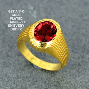 Può includere: Un anello placcato in oro con una grande gemma rossa ovale. L'anello ha una fascia strutturata con linee verticali e una lunetta decorativa. Il testo sull'immagine recita: GET A 14K GOLD PLATED CHAIN FREE ON EVERY ORDER.