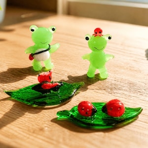Peut inclure: Deux petites figurines de grenouilles en verre vert, l'une avec une sangle noire et un sac blanc, l'autre avec une minuscule coccinelle rouge sur la tête. Deux feuilles en verre vert sont incluses, l'une avec une coccinelle et l'autre avec deux.