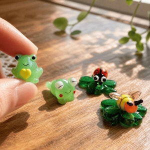Peut inclure: Une main tenant une petite figurine de grenouille verte translucide avec un cœur jaune. D'autres figurines incluent une grenouille endormie, une coccinelle et une abeille, le tout sur une surface en bois. Les figurines sont petites et détaillées.