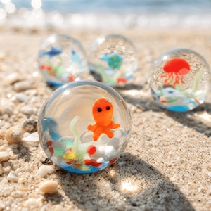 Könnte beinhalten: Vier klare Glaskugeln mit Miniatur-Meeresbewohnern. Die vordere Kugel zeigt einen orangefarbenen Oktopus mit schwarzen Augen, grünen Algen und weißen Kieselsteinen. Andere Kugeln enthalten eine rote Qualle, blaue Fische und grüne Wasserpflanzen.