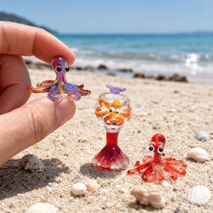 May include: Three miniature glass octopus figurines. One iridescent purple and orange octopus is held between fingers. A second orange octopus sits inside a clear glass goblet with a red base. A third solid red octopus rests on the sand.