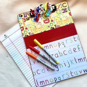 May include: A decorative clipboard with a yellow background featuring letters and school-themed illustrations. It has a red accent and a metal clip. Three Sharpie markers in yellow, red, and orange rest on a white alphabet board, with a lined paper pad.