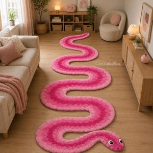 Puede incluir: Una alfombra larga y copetuda con forma de serpiente, que presenta varios tonos de rosa en un patrón degradado. La alfombra tiene una cabeza redondeada con dos ojos blancos y pupilas negras, extendiéndose por el suelo en una curva sinuosa.